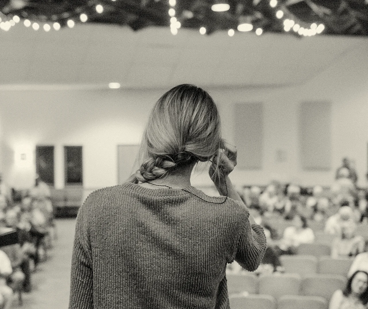 A girl stands on a stage with her back toward the camera, facing the crowd, tucking her hair behind her ear as the audience looks at her.